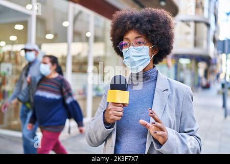 afroamerikanische Reporterin trägt eine medizinische Maske und benutzt ein Mikrofon, das auf der Straße arbeitet Stockfoto