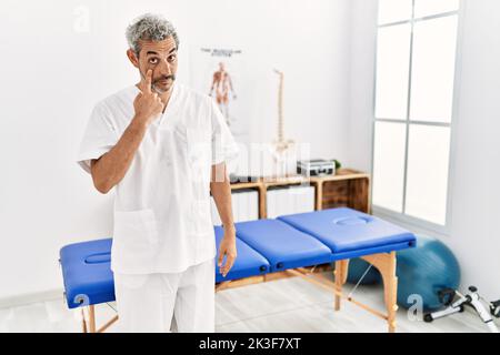 hispanischer Therapeut im mittleren Alter, der in der Schmerzklinik arbeitet und auf das Auge zeigt, während er Ihnen Geste, verdächtiger Ausdruck, zusieht Stockfoto