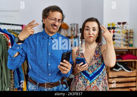 Interrassisches Paar im mittleren Alter im Einzelhandelsgeschäft, das Smartphone schießt und sich selbst tötet, indem es Hand und Finger an den Kopf zeigt wie eine Waffe, eine Selbstmordgeste Stockfoto