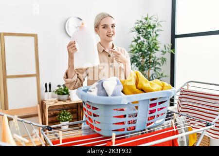 Junge kaukasische Frau, die ihre Wäsche waschen und die Waschmittelflasche lächelnd hält, zeigt glücklich mit Hand und Finger Stockfoto
