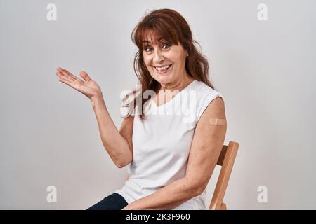 hispanische Frau mittleren Alters, die einen Impfstoff erhält, zeigt einen Arm mit einem Band, das zur Seite zeigt, mit offenen Händen, mit offenen Handflächen, die einen Kopierbereich zeigen und Werbung präsentieren Stockfoto