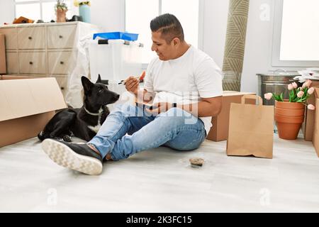 Junger latein, der mit Hund taka away Sushi isst, sitzt auf dem Boden in einem neuen Zuhause. Stockfoto