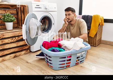 Junger gutaussehender Mann, der schmutzige Wäsche in die Waschmaschine legt und gestresst und nervös mit den Händen auf dem Mund beißt Nägel. Angstproblem. Stockfoto