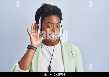 afroamerikanische Frau mit Callcenter-Agenten-Headset lächelt mit der Hand über dem Ohr und hört Gerüchten oder Klatsch zu. Taubheitskonzept. Stockfoto