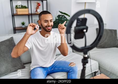 afroamerikanischer Mann, der Vlog-Tutorial über Bitcoin mit Smartphone zu Hause aufzeichnet, lächelt glücklich und zeigt mit Hand und Finger zur Seite Stockfoto