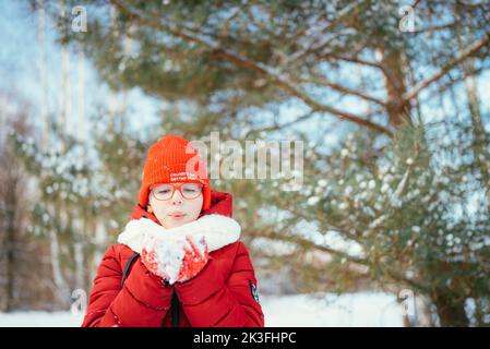 Portrait eines Mädchens in einem roten Hut und Schal, das an einem kalten Tag in einem verschneiten Wald auf dem Schnee weht. Stockfoto