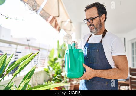 Mann mittleren Alters lächelt zuversichtlich Gießpflanzen auf der Terrasse zu Hause Stockfoto