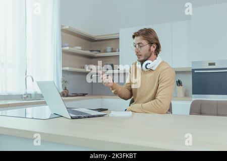 Ein junger Geschäftsmann, der von zu Hause aus in der Küche arbeitet, ein Mann, der einen Laptop für Videoanruf verwendet und sich auf Online-Meetings mit Kollegen konzentriert und ernsthaft darüber spricht. Stockfoto