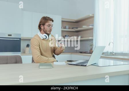 Ein junger Geschäftsmann, der von zu Hause aus in der Küche arbeitet, ein Mann, der einen Laptop für Videoanruf verwendet und sich auf Online-Meetings mit Kollegen konzentriert und ernsthaft darüber spricht. Stockfoto