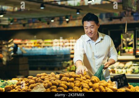 In einem Supermarkt in der Gemüseabteilung pflückt ein junger, gutaussehender asiatischer Mann Kartoffeln in einer grünen Plastiktüte. Stockfoto