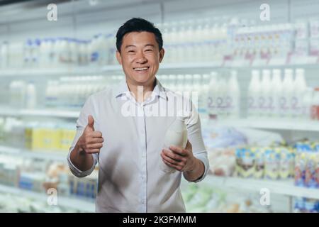 Werbung. Porträt eines jungen, hübschen asiatischen Mannes, der in einem Supermarkt in der Milchabteilung steht und eine Plastikflasche Milch in den Händen hält. Er zeigt auf OK, lächelt, schaut auf die Kamera. Stockfoto