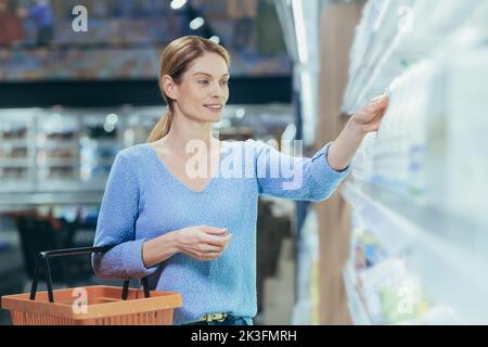 Eine junge, schöne Hausfrau steht mit einem Korb in der Hand in einem Supermarkt und wählt Milchprodukte aus. Er nimmt eine Flasche mit Milch, Joghurt, lächelt. Stockfoto