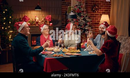 Fröhliche Familienmitglieder tragen festliche Hüte, während sie Gläser mit Champagner anklirren. Fröhliche, vielfältige Menschen lachen herzlich, während sie das Weihnachtsessen gemeinsam zu Hause genießen. Stockfoto