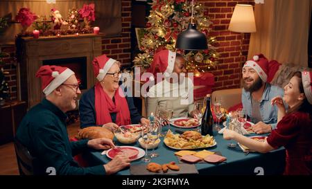 Fröhlich lachend, herzlich, multiethnische Familie, die Sekt toasten und beim Abendessen Weihnachtslieder singen. Festliche, vielfältige Menschen tragen traditionelle Hüte und genießen gemeinsam den Winterurlaub zu Hause. Stockfoto