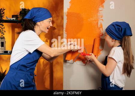 Fröhliche Menschen malen orange Wände zu Hause, mit Roller Pinsel und Farbe zu malen Wohnung zu schmücken. Frau mit jungen Kindern, die Spaß daran haben, gemeinsam zu Hause zu renovieren, Inneneinrichtung. Stockfoto