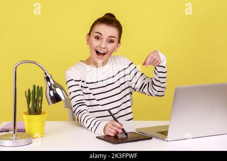 Entzückte kreative Designerin, die am Arbeitsplatz mit einem grafischen Tablet und einem Laptop sitzt und mit Spannung auf die Kamera zeigt. Studio-Innenaufnahme isoliert auf gelbem Hintergrund. Stockfoto