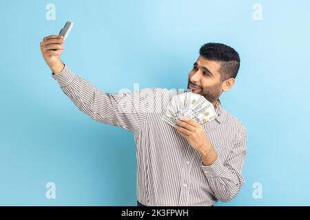 Äußerst zufriedener Geschäftsmann macht Selfie-Foto mit Haufen Dollar auf dem Smartphone, prahlte mit Reichtum, trägt gestreiftes Hemd. Innenaufnahme des Studios isoliert auf blauem Hintergrund. Stockfoto