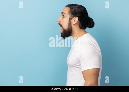 Die Seitenansicht eines überraschten Mannes mit Bart, der ein weißes T-Shirt trägt und schockiert aussieht, sieht etwas Erstaunliches. Innenaufnahme des Studios isoliert auf blauem Hintergrund. Stockfoto