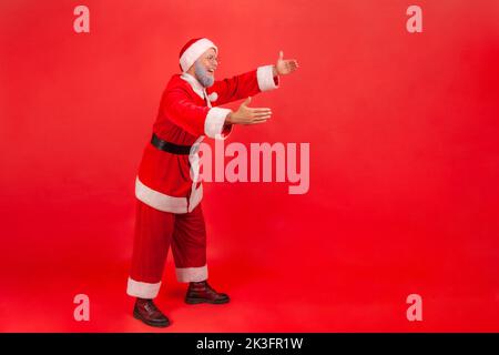 Ganzkörperaufnahme eines älteren Mannes mit grauem Bart, der das weihnachtsmann-Kostüm trägt und die Hände ausstreckt, um jemanden zu umarmen. Innenaufnahme des Studios isoliert auf rotem Hintergrund. Stockfoto