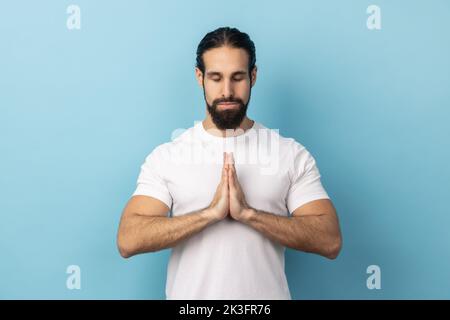 Mann mit Bart trägt weißes T-Shirt konzentriert seinen Geist, halten die Hände namaste Geste, meditieren, Yoga-Übung Atemtechnik reduzieren Stress. Innenaufnahme des Studios isoliert auf blauem Hintergrund. Stockfoto
