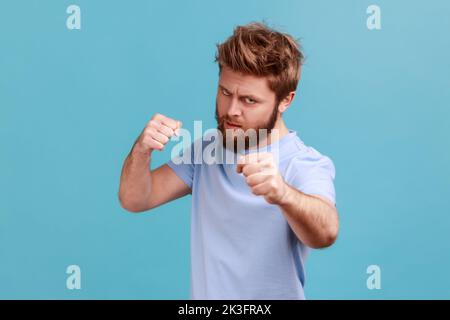 Porträt eines bärtigen Mannes, der mit Boxfäusten und bereit zum Angriff oder zur Verteidigung steht, mit wütendem Gesicht schaut, mit Aggression auf die Kamera blickt. Innenaufnahme des Studios isoliert auf blauem Hintergrund. Stockfoto