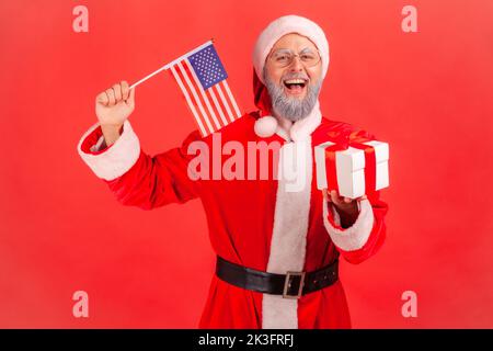Porträt eines aufgeregten älteren Mannes mit grauem Bart, der das weihnachtsmann-Kostüm trägt und die USA-Flagge in den Händen hält und eine Geschenkiste für die Winterferien gibt. Innenaufnahme des Studios isoliert auf rotem Hintergrund. Stockfoto