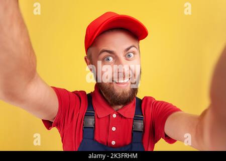 Optimistischer glücklicher Mann mit roter Mütze und blauer Uniform, der Selfie macht, auf die Kamera schaut lustiger Gesichtsausdruck POV, Standpunkt des Fotos. Innenaufnahme des Studios isoliert auf gelbem Hintergrund. Stockfoto