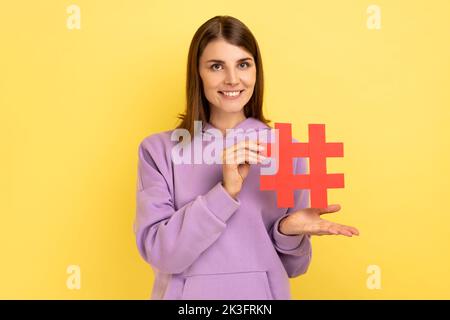 Porträt einer jungen Frau, die im Stehen steht und roten Hashtag präsentiert, Blog-Trends markiert, virales Thema in sozialen Netzwerken, purpurfarbenes Kapuzenpullover trägt. Innenaufnahme des Studios isoliert auf gelbem Hintergrund. Stockfoto