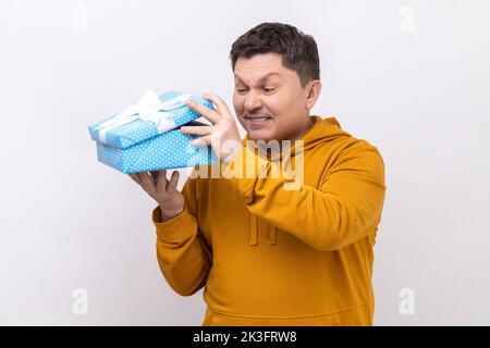 Porträt eines Mannes mittleren Alters, der eine Geschenkbox öffnet und nach innen guckt, mit runzelndem Gesicht, unangenehmer Überraschung, mit einem Hoodie im urbanen Stil. Innenaufnahme des Studios isoliert auf weißem Hintergrund. Stockfoto