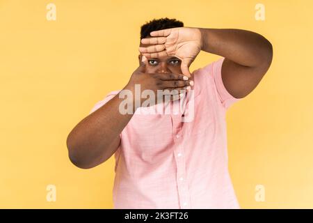 Halten Sie Momente fest. Porträt eines Mannes in einem rosa Hemd, der mit den Fingern durch die Form eines Fotorahmens schaut, in der Ferne sieht, die Kamera fokussiert und zoomt. Innenaufnahme des Studios isoliert auf gelbem Hintergrund. Stockfoto