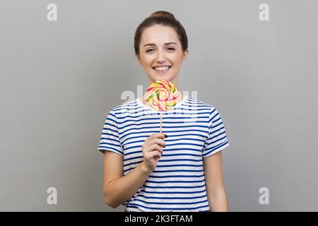 Porträt einer lächelnden Frau, die ein gestreiftes T-Shirt trägt und einen gestreiften bunten Lutscher auf dem Stock in der Hand hält, verrückt nach süßer Süßigkeit. Innenaufnahme des Studios isoliert auf grauem Hintergrund. Stockfoto