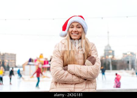Outdoor-Porträt junge schöne modische Frau trägt Santa Hut, posiert Straße der europäischen Stadt. Winter Weihnachtsferien Konzept. Eislaufbahn Hintergrund. Kaukasische weibliche Winterkleidung Stockfoto