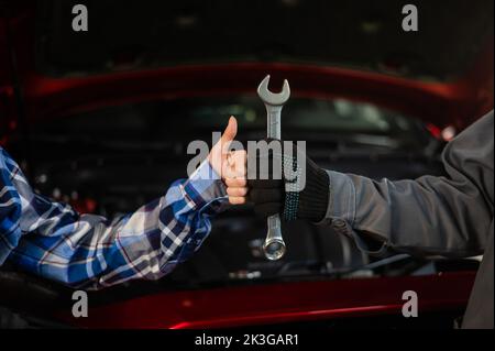 Nahaufnahme des Daumens der Frau und der Hand des Mechanikers mit Werkzeug. Automeister in Handschuhen zeigt nach Beendigung der Arbeit eine positive Geste Stockfoto