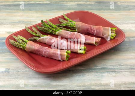 Rohe Spargelbüschen, die in Prosciutto in Scheiben geschnitten und auf einer roten Platte zum Kochen eingewickelt sind Stockfoto