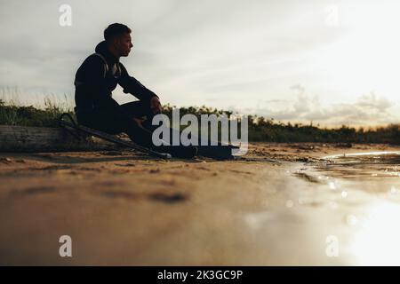 Junger männlicher Taucher, der am Strand mit einer Speergun und Speerfischerausrüstung sitzt. Abenteuerlicher junger Mann, der den Sonnenuntergang nachdenklich betrachtet. Stockfoto