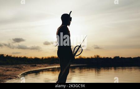 Seitenansicht des Speerfischers, der am Ufer des Meeres steht und ins Meer blickt. Männlicher Taucher geht Speerfischen mit einer Speergun.