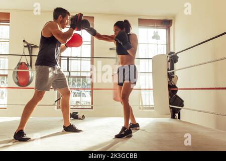 Männlicher Trainer hilft einer jungen Boxerin mit umgekehrten Schlägen in einem Boxring. Boxtrainer trainiert eine sportliche junge Frau im Fitnessstudio. Stockfoto