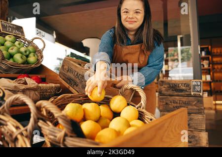 Fröhliche Frau mit Down-Syndrom, die frische Produkte in einem Delikatessengeschäft auffüllt. Eine Frau mit geistiger Behinderung, die in einem lokalen Supermarkt arbeitet. Stockfoto