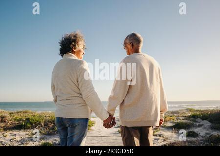 Rückansicht eines glücklichen älteren Paares, das sich die Hände hielt, während er eine Fußgängerbrücke am Strand entlang ging. Ein romantisches, älteres Paar, das einen erfrischenden Urlaub am Meer nimmt Stockfoto