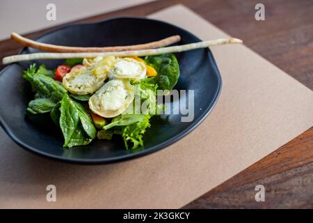 Frühstück am Morgen, cremiges Omelette und Buratta Chesese, Salat Avocado und grüne Blätter auf schwarzem Teller.Essen gesundes Konzept.Salat mit Tomaten Stockfoto
