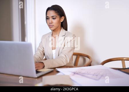 Online kreativ werden. Eine junge Frau tippt auf ihrem Laptop, während Skizzen und Bleistifte neben ihr auf dem Schreibtisch liegen. Stockfoto