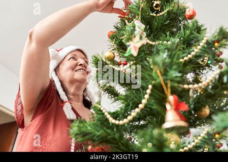 Fröhliche ältere lateinerin, die lächelt, während sie ihren Weihnachtsbaum zu Hause schmückt und eine rote Weihnachtsmann-Mütze mit weißen Zöpfen trägt. Die Freude des Heiligen Stockfoto