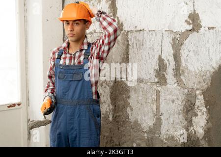 Nachdenklicher junger Baumeister mit einem Spachtel auf dem Hintergrund einer Wand aus weißen Ziegeln, eine Wand aus Betonmörtel, ein Lieblingsberuf, Haus r Stockfoto