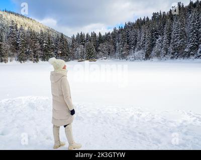 Frau in weißen Kleidern genießt gefrorenen See Synevyr und Wald in den Karpaten Berge. Wintersee zwischen den schneebedeckten Wäldern. Wunderschön Stockfoto