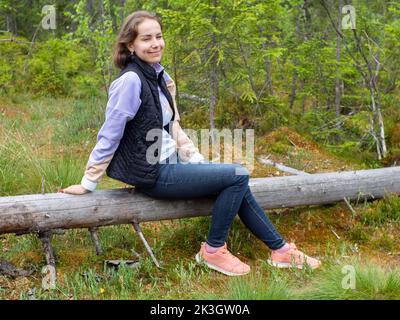 Wanderer, der während eines Waldspaziergangs Pause einnahm. Junge kaukasische Frau, die in einem grünen Wald auf einem umgestürzten Baum sitzt. Nature Connection-Konzept. Speicherplatz kopieren. Eine Tour Stockfoto