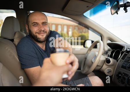 Porträt eines Mannes, der im Auto sitzt und Kaffee für unterwegs kauft. Mann, der sein Auto fährt und Kaffee hält, während er Freude daran atmt Stockfoto