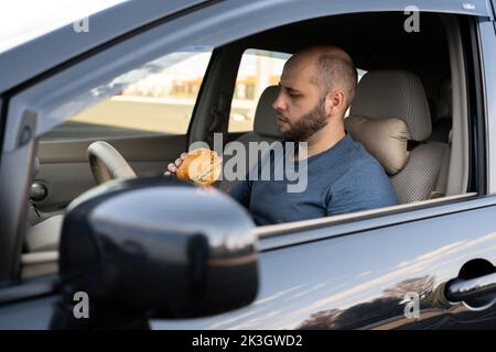 Fahrer Mann am Steuer beim Essen im Verkehr, essen Hamburger im Auto. Stadtleben. Stockfoto