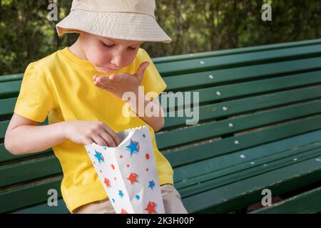 Junge in der Sommerkleidung auf der Parkbank isst Popcorn, Sommernahrung, Snack, Süßigkeiten. Stockfoto