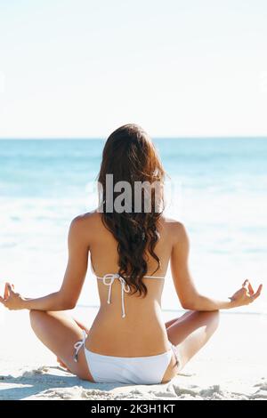 Frieden von der Natur inspiriert. Eine junge Frau, die in der Sommersonne am Strand eine Yoga-Routine durchführt - Rückansicht. Stockfoto