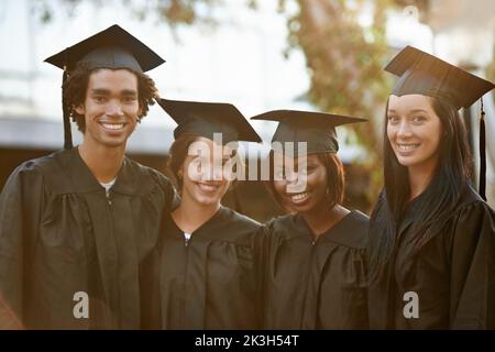 Stolz darauf, Absolventen zu sein. Eine Gruppe lächelnder Hochschulabsolventen, die in Mütze und Kleid zusammenstehen. Stockfoto